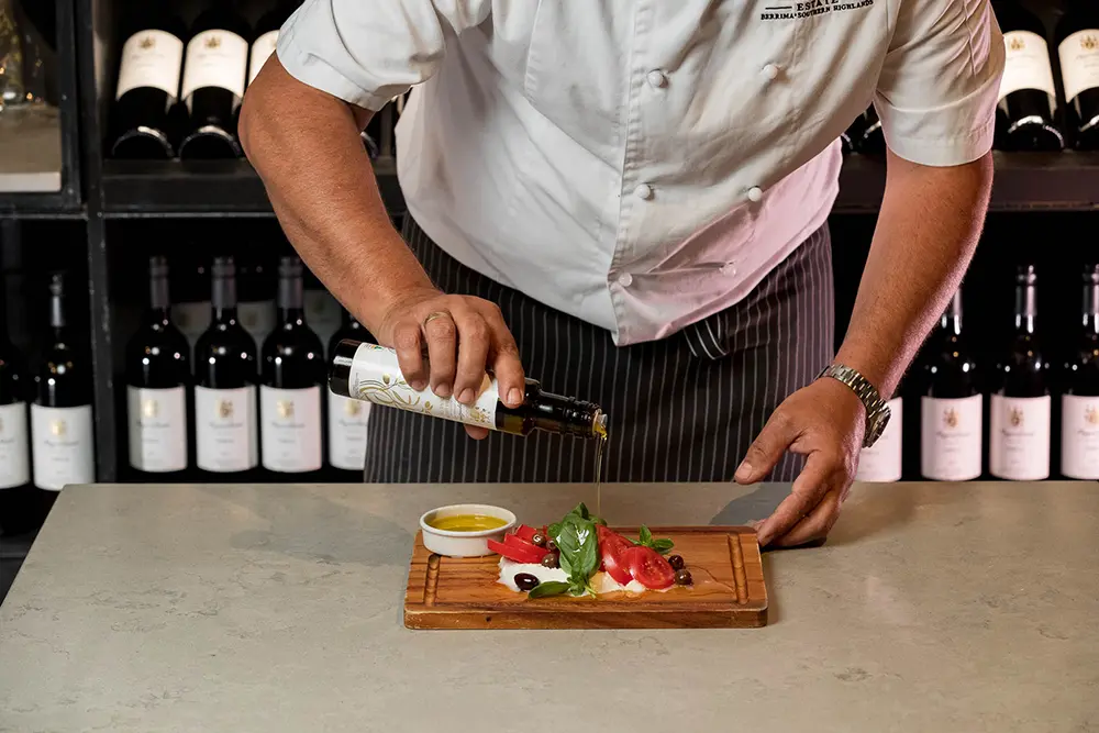 Chef drizzling high-quality extra virgin olive oil over a fresh tomato and mozzarella salad, enhancing flavor and nutrition in a professional kitchen setting.