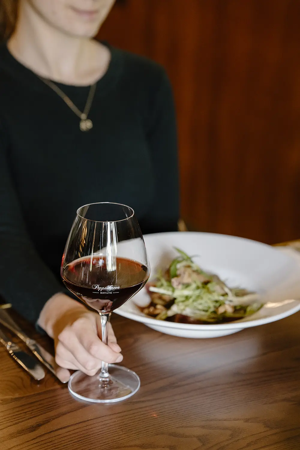 A woman enjoying a glass of red wine alongside a dish of garlic olive oil pasta, highlighting a refined dining experience with freshly prepared Italian cuisine made with high-quality extra virgin olive oil.