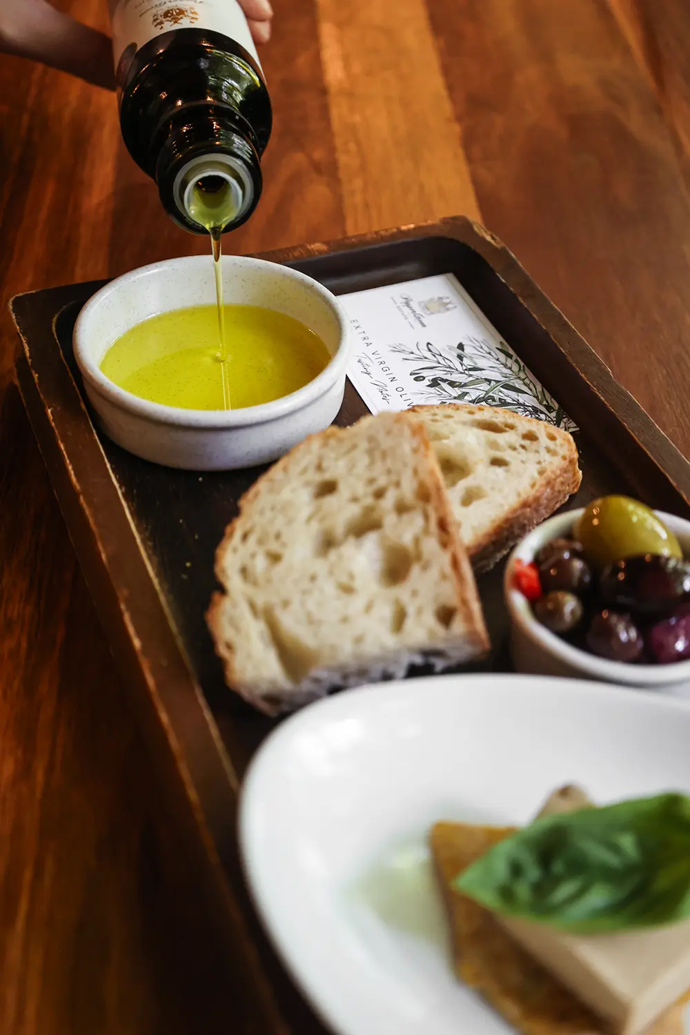 Pouring extra virgin olive oil into a dish with rustic bread, olives, and cheese, showcasing the best ways to enjoy olive oil with salads, seafood, and Mediterranean cuisine.