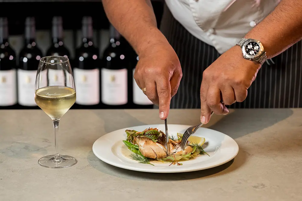 Chef slicing seared salmon fillet garnished with herbs, paired with white wine, highlighting the use of premium olive oil and fresh lemon garlic sauce for a refined culinary experience.