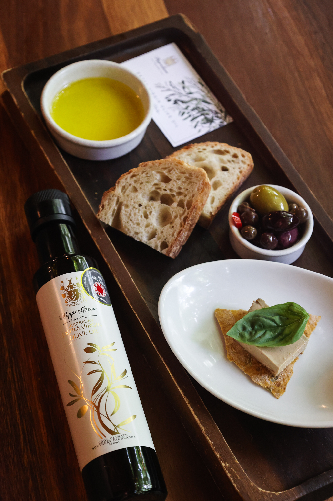 A bottle of PepperGreen Estate extra virgin olive oil served with bread, tofu appetizer and olives, showcasing its versatility for healthy stir-fry, dipping, and Mediterranean-style meals