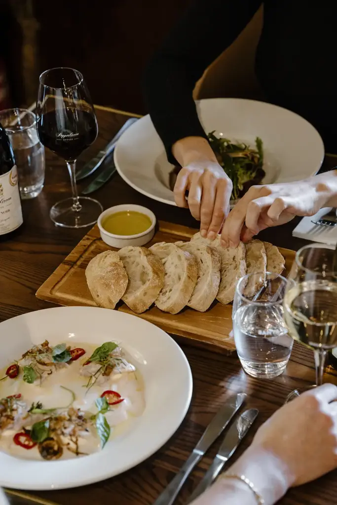 Hands reaching for sliced bread on a wooden dining table with an olive oil bottle, wine glasses, salad, and fish.